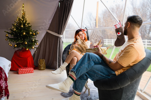 A young couple is sitting in cozy glamping. The man gives a gift to the woman for Christmas.