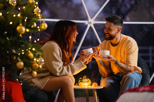 A young couple is sitting at a table by a huge window talking and drinking tea from large cups. Evening. Christmas tree.