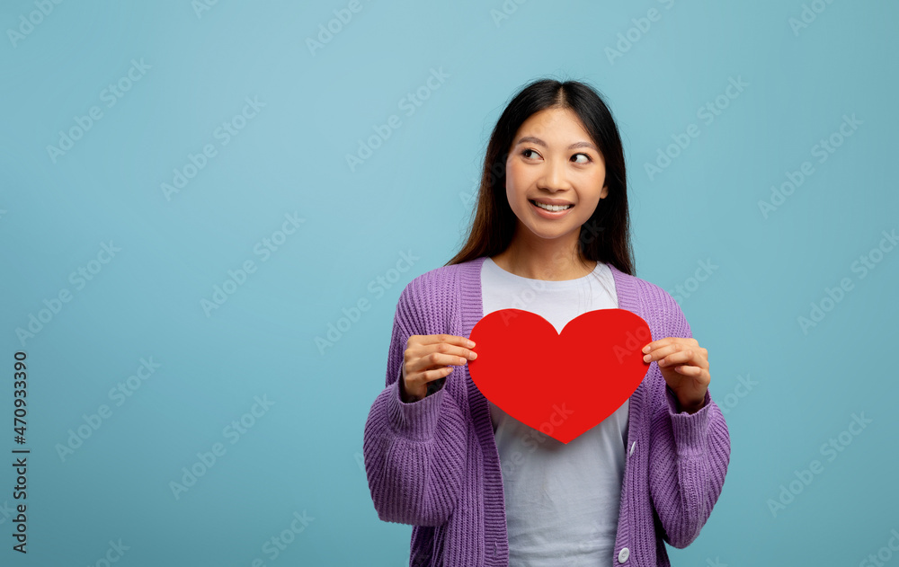 St. Valentines day concept. Playful asian lady holding paper heart and looking aside at free space on blue background St. Valentines day concept. Playful asian lady holding paper heart and looking aside at free space on blue background