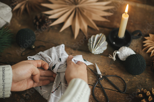Zero waste christmas furoshiki gift. Hands wrapping christmas gift box in modern fabric on rustic wooden table with scissors, craft paper star, candle. Atmospheric moody time, nordic style photo
