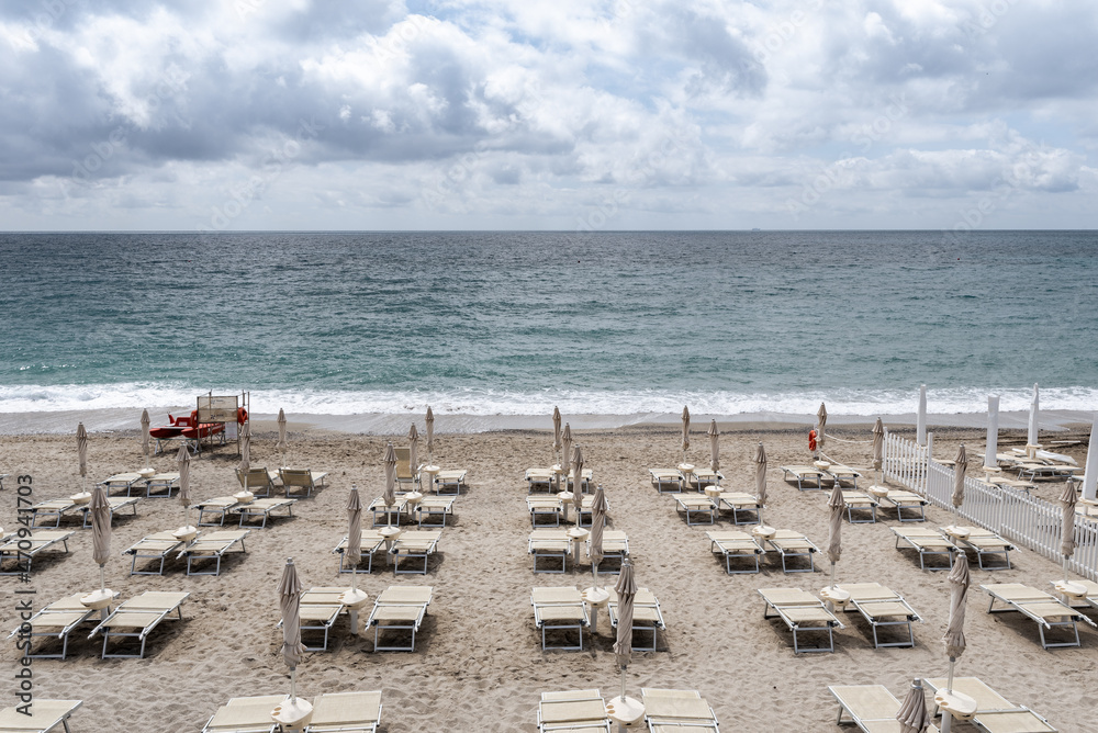 Finale Ligure. May 21, 2021. View from the Lungomare Italia street on an empty stretch of beach with white sunbeds and umbrellas and a lifeguard spot with a small rescue boat, on a cloudy day.