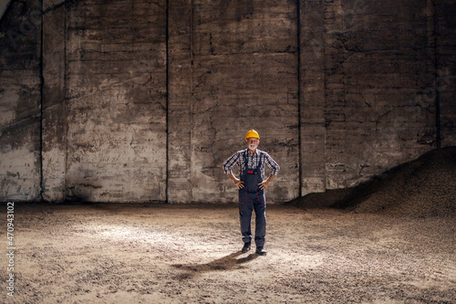 An old hardworking worker in overalls standing with hands on hips in the dark industrial room full of sugar beet products. Sugar factory, production of sweets © dusanpetkovic1