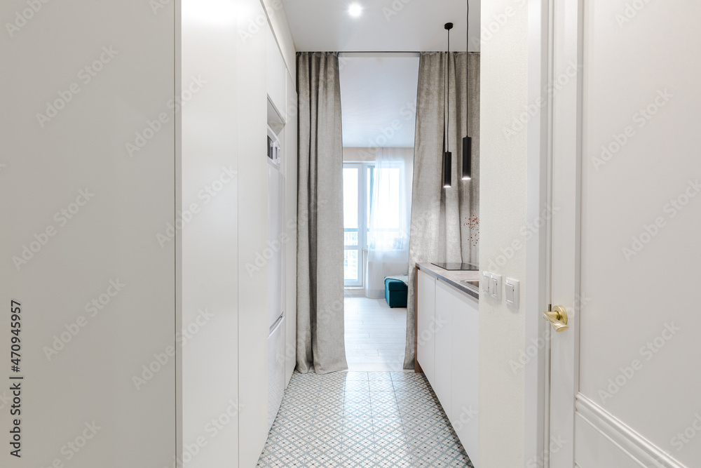Minimalistic design of a white studio apartment. View from the entrance hall to a compact built-in kitchen, a cozy room with a sofa and a round dining table Minimalistic design of a white studio apartment. View from the entrance hall to a compact built-in kitchen, a cozy room with a sofa and a round dining table