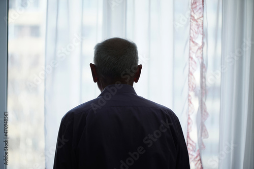 Silhouette of retired man looking through window with transparent curtain