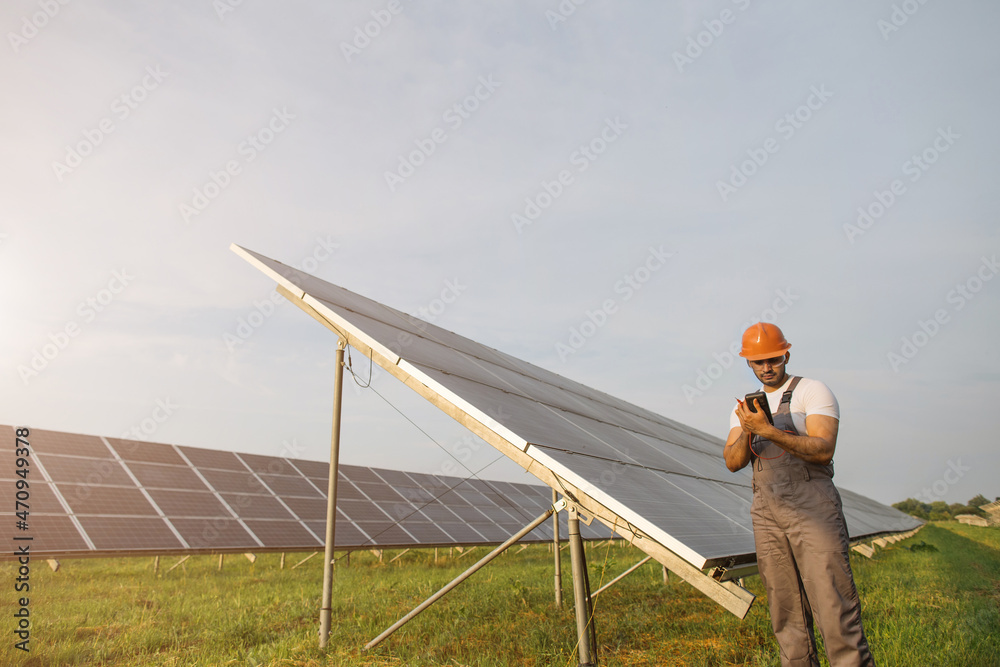 Indian worker in uniform, safety helmet and glasses using multimeter while examining work of solar panels. Concept of people, maintenance and green energy.