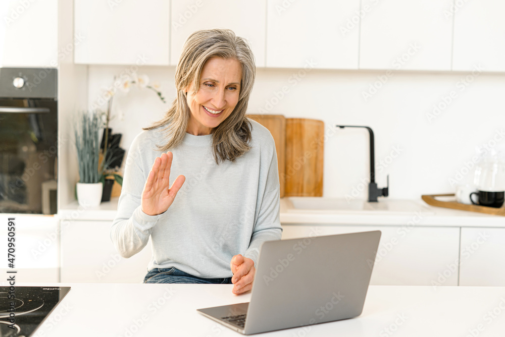 Good-looking senior mature woman is using laptop for video connection, making a call, looking at computer screen and waving hand. Gray-haired grandmother in touch with family Good-looking senior mature woman is using laptop for video connection, making a call, looking at computer screen and waving hand. Gray-haired grandmother in touch with family