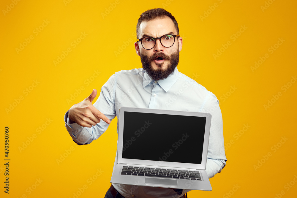 Bearded male wearing beige shirt and eyeglasses pointing at new laptop in hands on yellow background. 