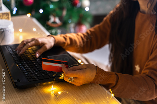 Christmas sales. Woman shopping with credit card in hand, in front of the laptop inside the house, near the Christmas tree. Vacation planning.