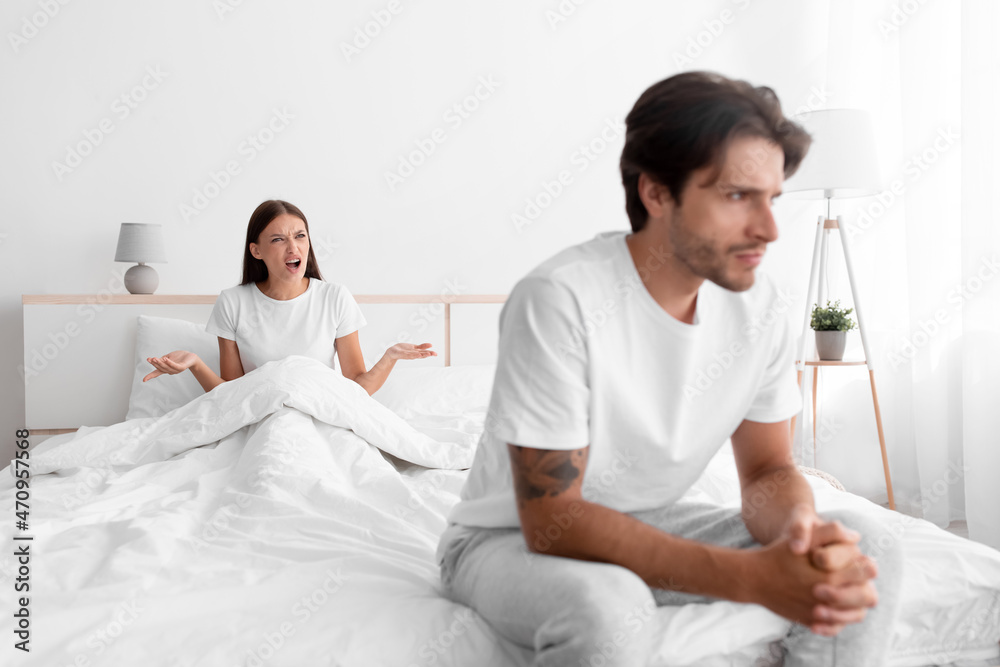 Unhappy frustration european young man ignoring angry woman, lady sits in bed and scream on guy in white bedroom Unhappy frustration european young man ignoring angry woman, lady sits in bed and scream on guy in white bedroom