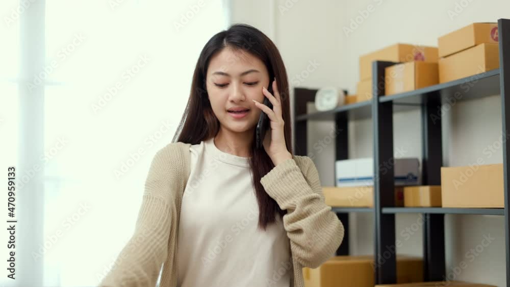 Busy Young asian business woman using mobile phone call receiving purchase order and check product on stock near the package shelf on background. Ecommerce business and shopping online concept. Busy Young asian business woman using mobile phone call receiving purchase order and check product on stock near the package shelf on background. Ecommerce business and shopping online concept.