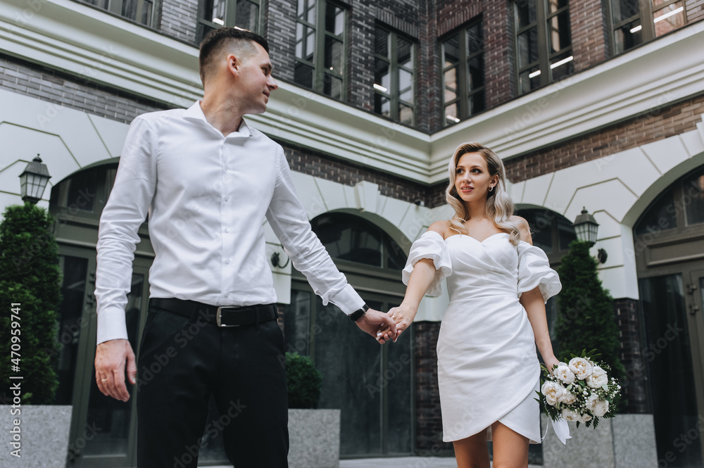 Stylish groom and a beautiful, young, curly-haired blonde bride in a white dress with a bouquet are walking holding hands against the backdrop of the city and buildings. Wedding photography. Stylish groom and a beautiful, young, curly-haired blonde bride in a white dress with a bouquet are walking holding hands against the backdrop of the city and buildings. Wedding photography.