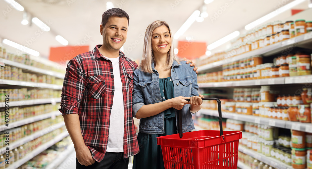 Young casual couple with a shopping basket smiling at camera and posing in a supermarket