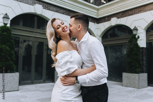 Stylish, smiling groom and a beautiful, young blonde bride in a white dress are embracing against the backdrop of the city and buildings. Wedding photography of the newlyweds, portrait.