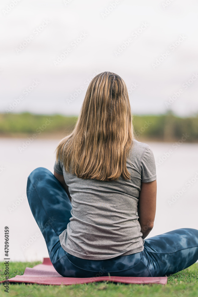 Back of a fit blonde woman sitting on a yoga mat facing a lake