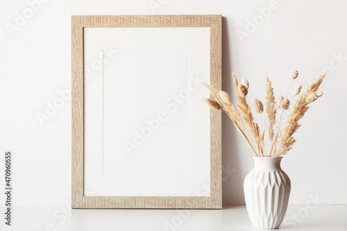 Empty wooden frame stands on white table with dried flowers in vase. Mockup poster frame close up in neutral color, minimalist interior.