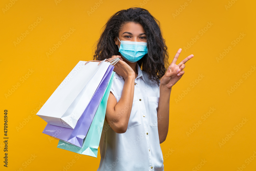 Portrait of a girl wearing protective medical mask on her face, posing with shopper bags and making v-sign gesture, isolated on yellow background. Secure shopping during the quarantine concept Portrait of a girl wearing protective medical mask on her face, posing with shopper bags and making v-sign gesture, isolated on yellow background. Secure shopping during the quarantine concept