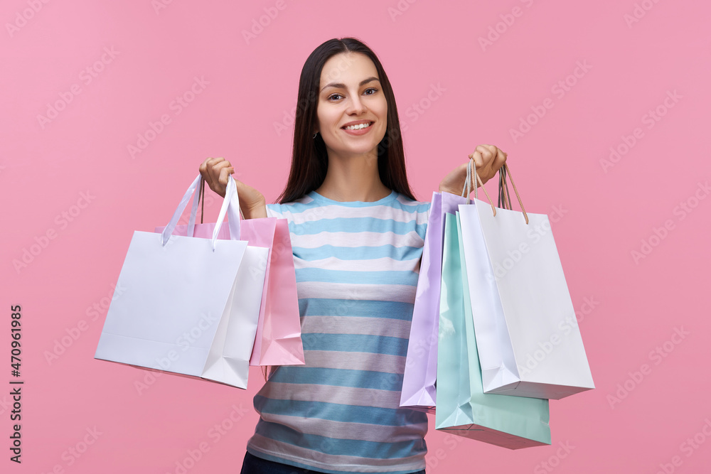 Portrait of casually dressed beautiful smiling brunette young woman posing with bunches of paper shopping bags in hands, isolated over pastel pink background Portrait of casually dressed beautiful smiling brunette young woman posing with bunches of paper shopping bags in hands, isolated over pastel pink background