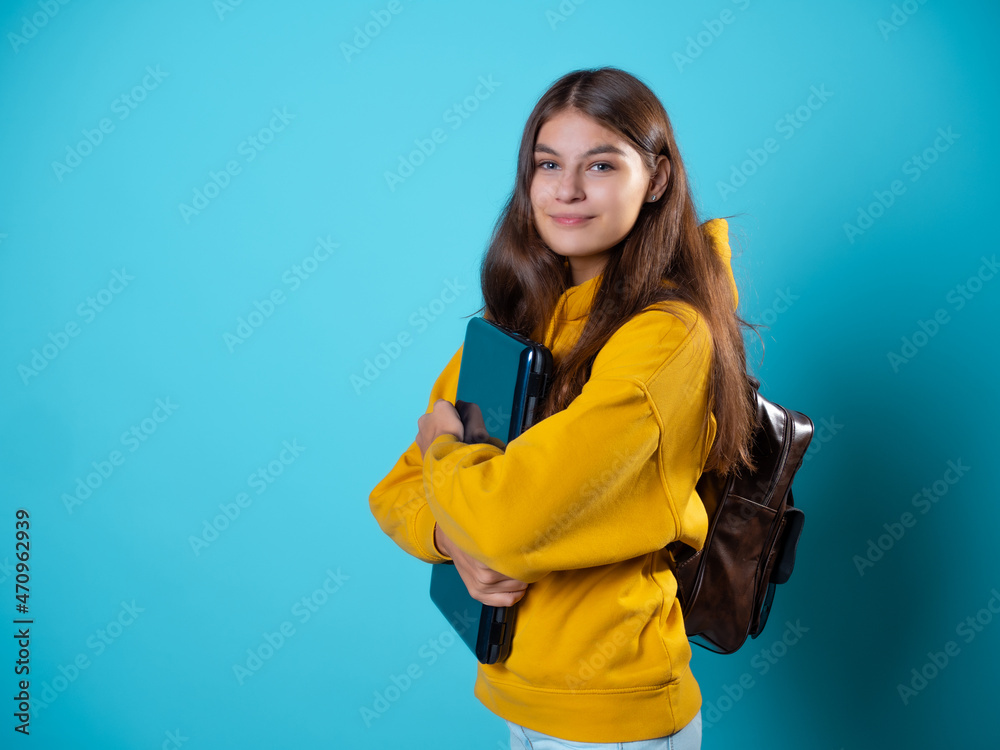 Work and study remotely, a new normal, a concept. A student with a laptop in her hands, a young brunette in a yellow hoodie on a blue background Work and study remotely, a new normal, a concept. A student with a laptop in her hands, a young brunette in a yellow hoodie on a blue background