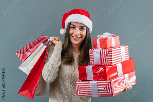 Portrait of happy smilig attractive young woman in Santa's hat and white knitted sweater, holding pile of gift boxes and shopping bags in hands photo