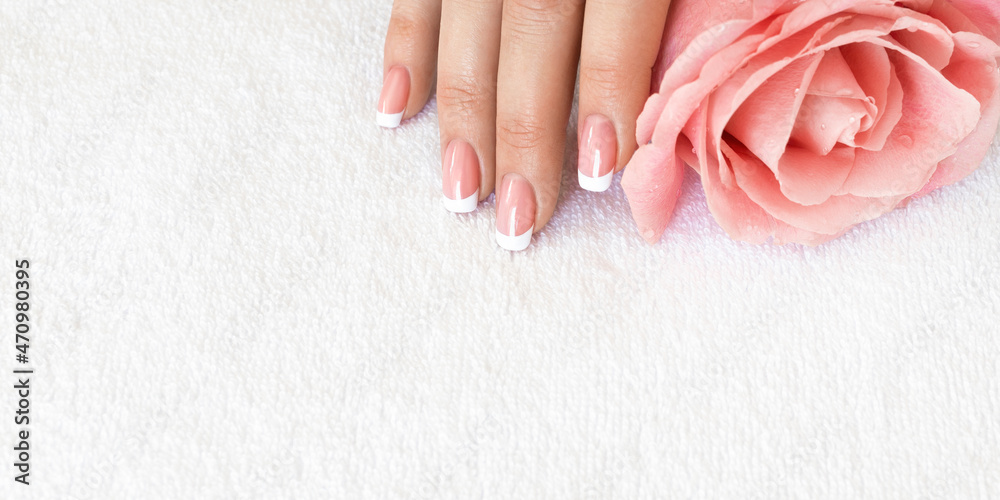 Naklejka premium Female fingers with beautiful French tips manicure on white terry towel and a soft pink rose flower, close up, selective focus.