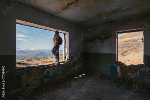 Woman motorcyclist in travel standing on abandoned building window enjoying mountain valley view with amazing snowy peaks skyline