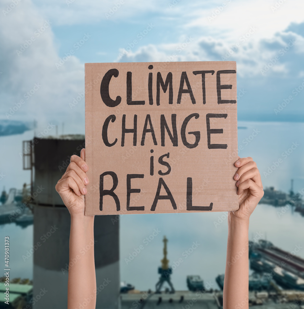 Protestor holding placard with text Climate Change Is Real and blurred view of industrial factory on background Protestor holding placard with text Climate Change Is Real and blurred view of industrial factory on background