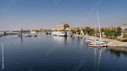Boats and feluccas float on the smooth blue water of the Nile. There are ships at the shore. Reflection. City houses are visible in the distance. Blue sky. Egypt. Aswan.