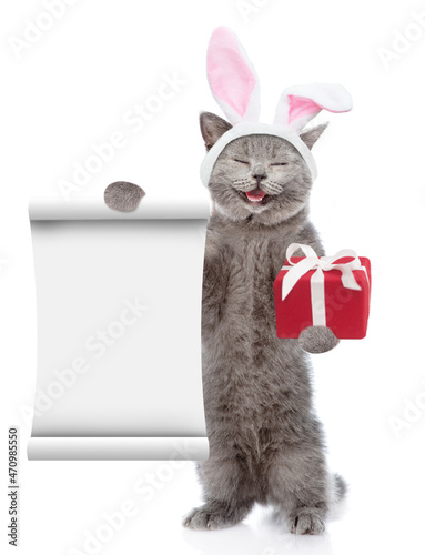 Happy kitten wearing Easter rabbits ears shows an empty list and holds gift box. Isolated on white background