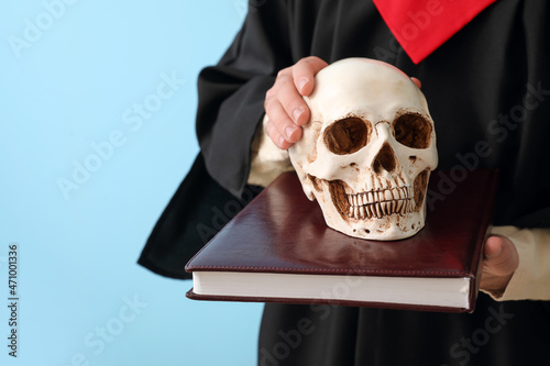 Male graduate with human skull and book on blue background, closeup © Pixel-Shot