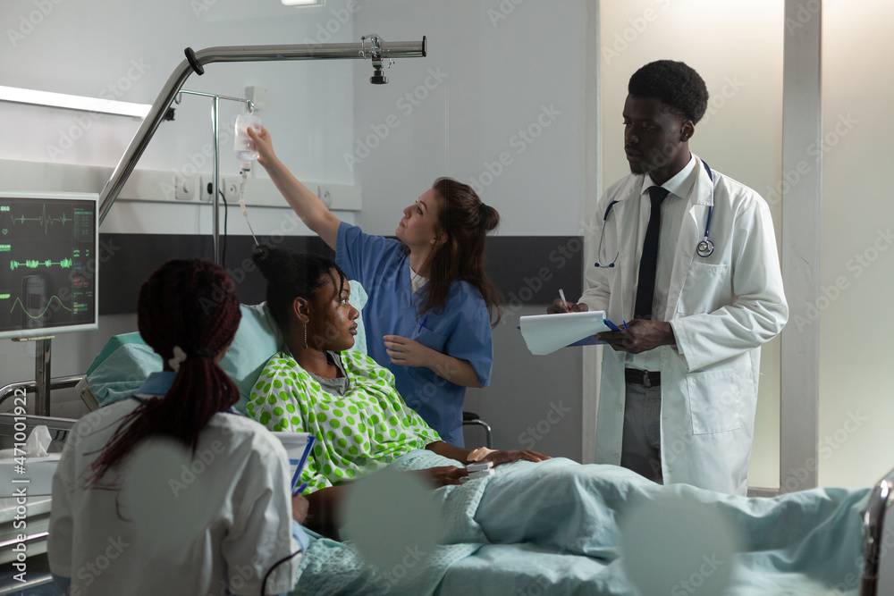 Woman assistant checking iv drip bag while african american therapist doctor analyzing sickness symptoms discussing medication treatment. Sick patient resting in bed recovering after surgery Woman assistant checking iv drip bag while african american therapist doctor analyzing sickness symptoms discussing medication treatment. Sick patient resting in bed recovering after surgery