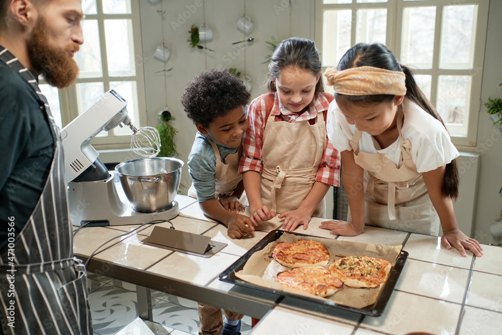Group of children in aprons looking at baked pizza on the tray and smiling, they cooking together with the chef Group of children in aprons looking at baked pizza on the tray and smiling, they cooking together with the chef