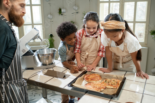Group of children in aprons looking at baked pizza on the tray and smiling, they cooking together with the chef