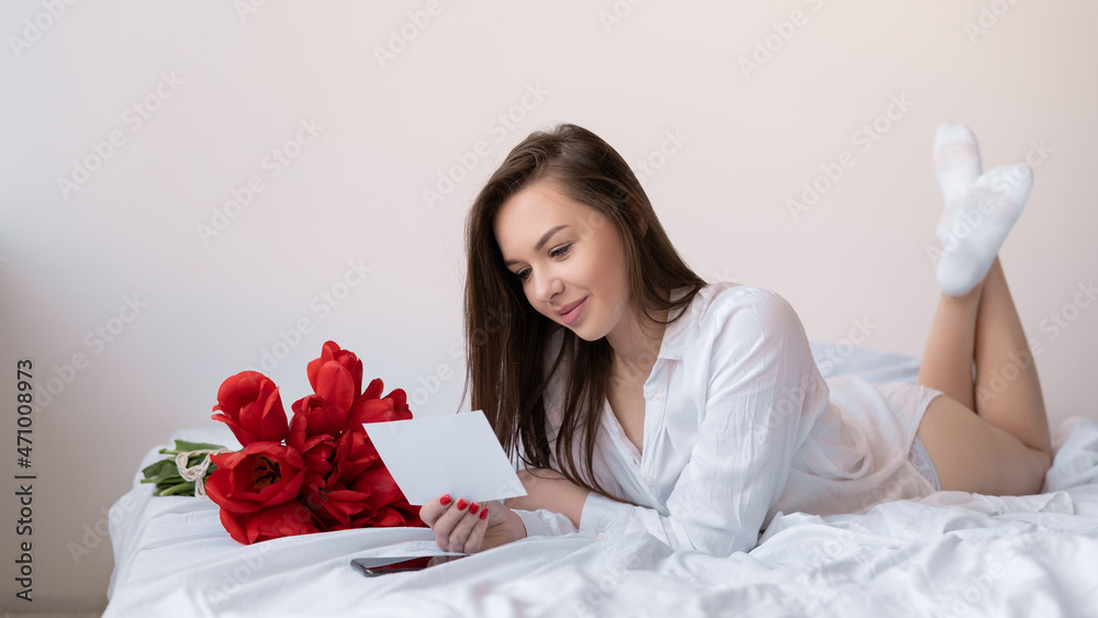 young beautiful caucasian woman lies on the bed near a bouquet of tulips reads a note. gift concept for birthday, valentine's day, anniversary. young beautiful caucasian woman lies on the bed near a bouquet of tulips reads a note. gift concept for birthday, valentine's day, anniversary.
