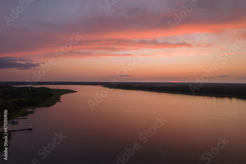 Aerial landscape of the lake surrounded by the green forest. Pastel orange clouds during summer sunset.