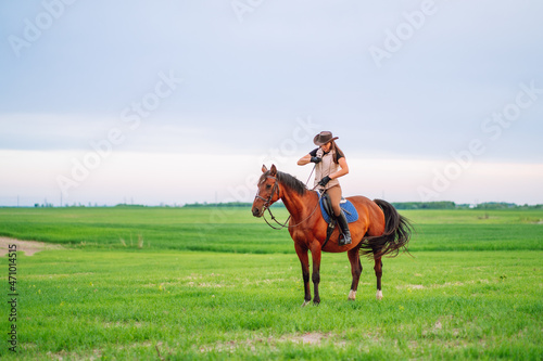 Young woman riding on brown horse in green field with blue sky on background. Horseback riding. Space for text
