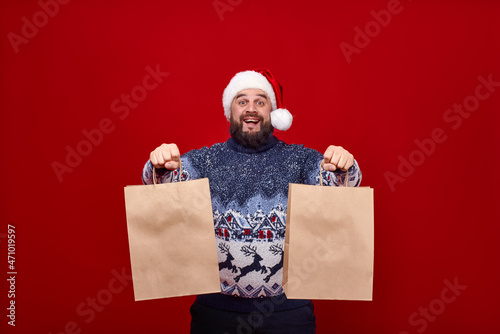 An emotional man in a Santa hat holds out two craft packages to the camera on a red background. A bearded guy in a sweater with deer. Place for the logo on brown paper bags.