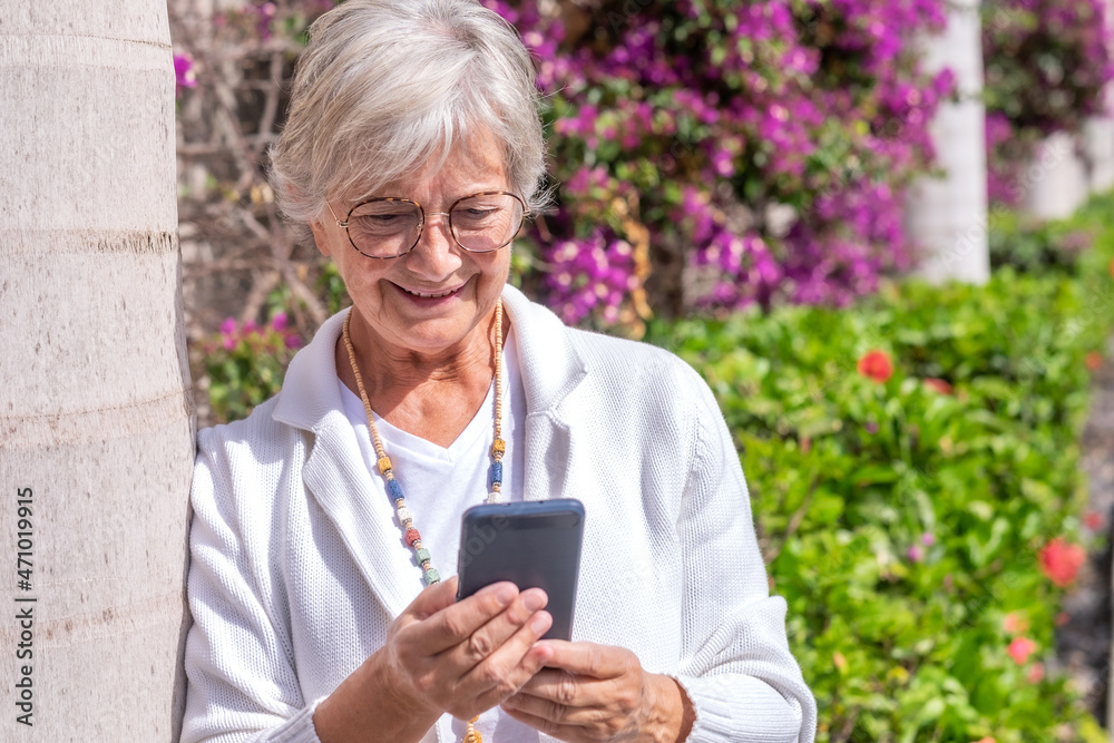Portrait of an elderly woman leaning on a plant while using the smart phone in a flowery avenue Portrait of an elderly woman leaning on a plant while using the smart phone in a flowery avenue