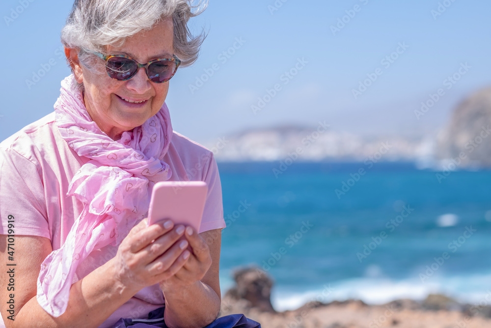 Smiling elderly woman with pink scarf using mobile phone in outdoor at sea. Horizon over water