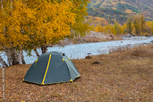 Green tent near river with colorful mountain forest in autumnseason. Autumn nature landscape. Tourism, trekking in fall season concept. Nature of Dzungarian Alatau in Kazakhstan.