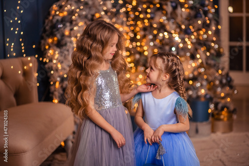 two elegant girls in beautiful dresses give each other a gift against the background of a Christmas tree with lights