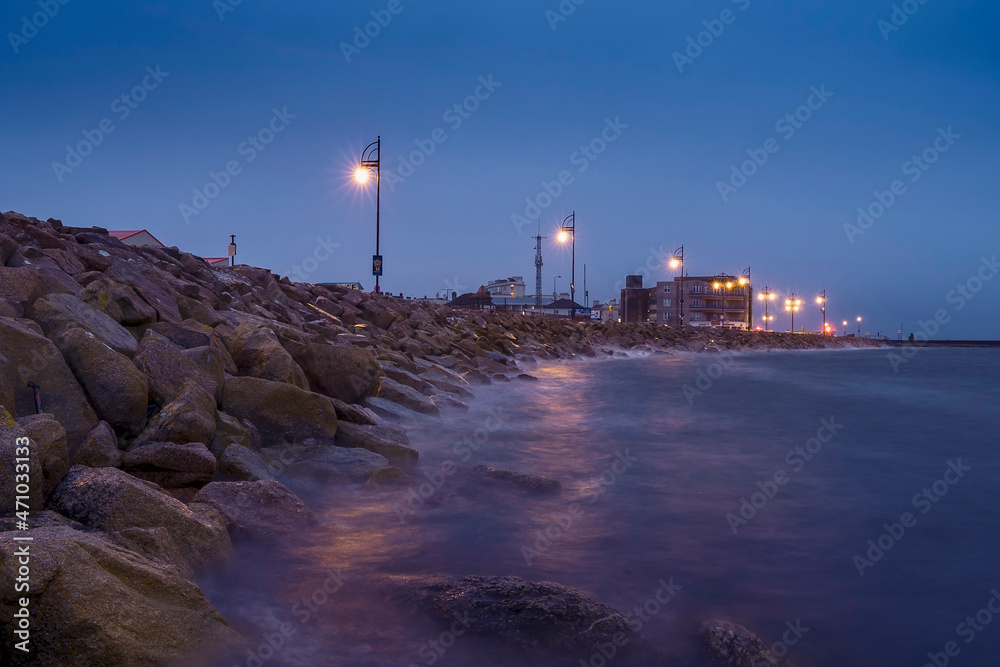 Rough stone coast of Salthill, Galway city Ireland. Blue hour. Town illumination reflects in calm water of the ocean. Calm and peaceful scene.