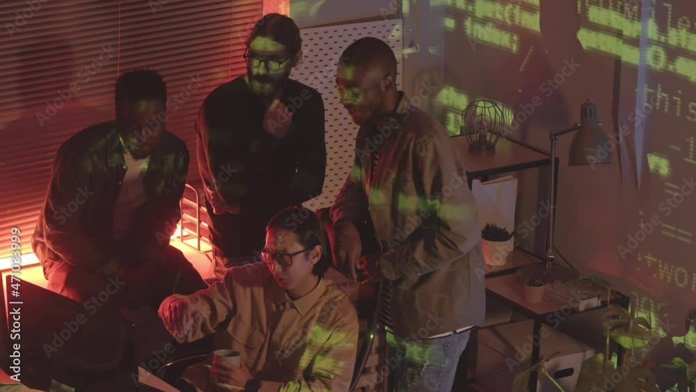 From-above slowmo shot of diverse team of programmers coworking at dark office with running green programming code lines projection From-above slowmo shot of diverse team of programmers coworking at dark office with running green programming code lines projection