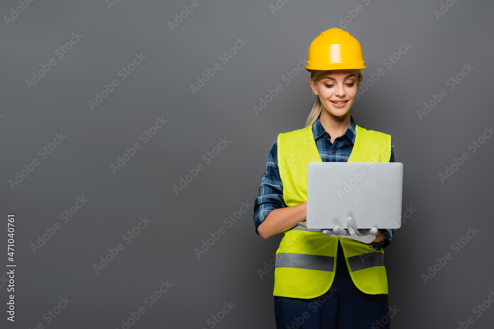 Positive builder in hardhat and protective vest using laptop isolated on grey Positive builder in hardhat and protective vest using laptop isolated on grey