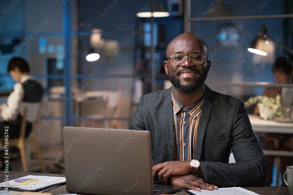 Portrait of African businessman smiling at camera while sitting at his workplace with laptop at office and working till night Portrait of African businessman smiling at camera while sitting at his workplace with laptop at office and working till night