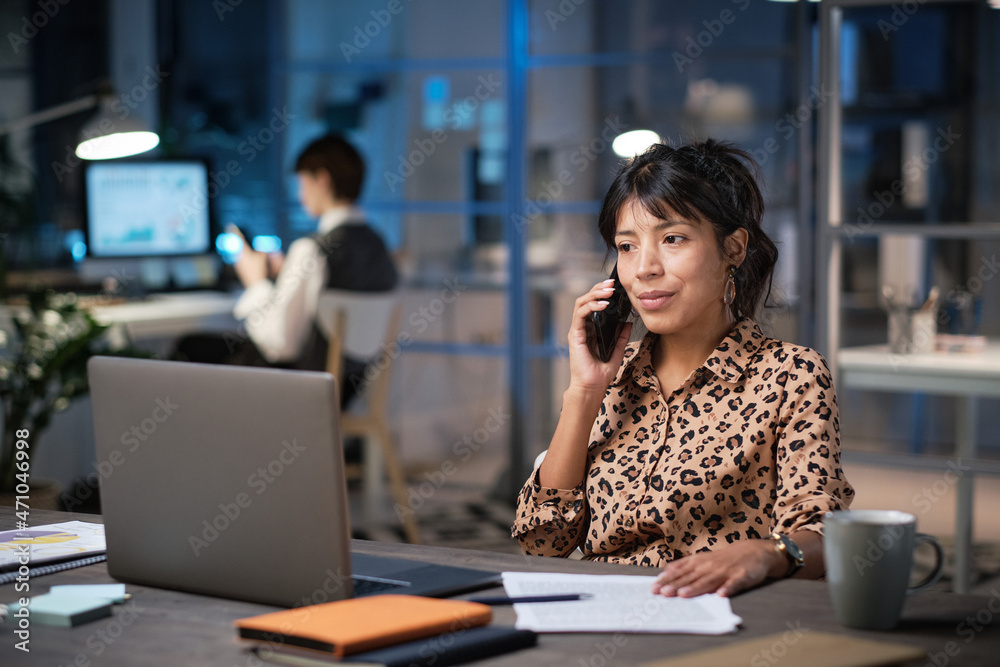 Mature businesswoman sitting at her workplace with documents looking at laptop screen and talking on the phone while working at office in the evening Mature businesswoman sitting at her workplace with documents looking at laptop screen and talking on the phone while working at office in the evening