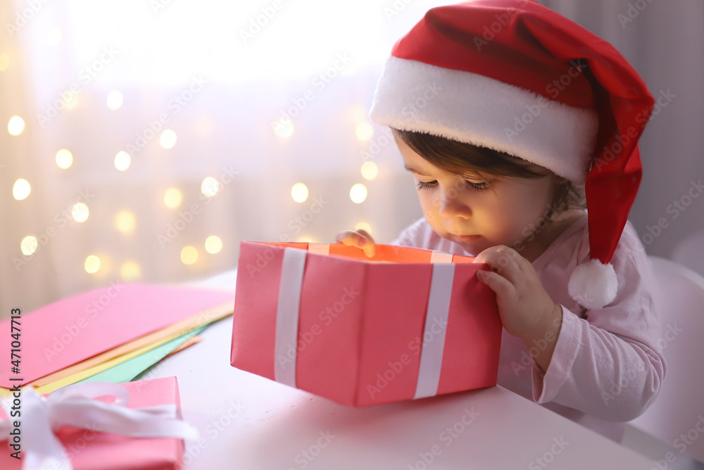 Little Christmas girl in a Santa hat looking into a gift box. Curious child with a New Year's gift. The background is defocused light. Concept of Christmas and New Year 2022. High quality photo Little Christmas girl in a Santa hat looking into a gift box. Curious child with a New Year's gift. The background is defocused light. Concept of Christmas and New Year 2022. High quality photo