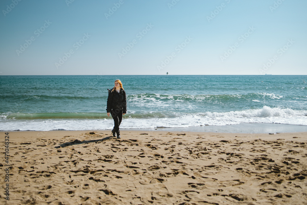 Blond girl in dark clothes with backpack walking on the winter beach