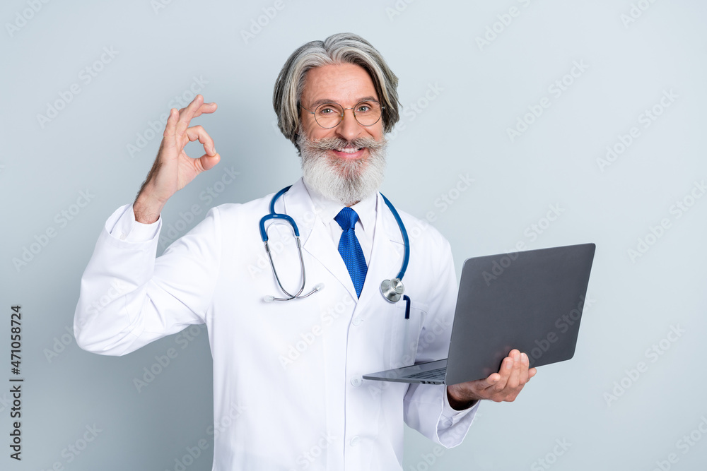 Photo of senior man cardiologist consultation use laptop show fingers okey symbol perfect ad isolated over grey color background Photo of senior man cardiologist consultation use laptop show fingers okey symbol perfect ad isolated over grey color background