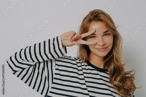 Happy young European woman makes peace disco gesture over eye v sign wears casual striped jumper