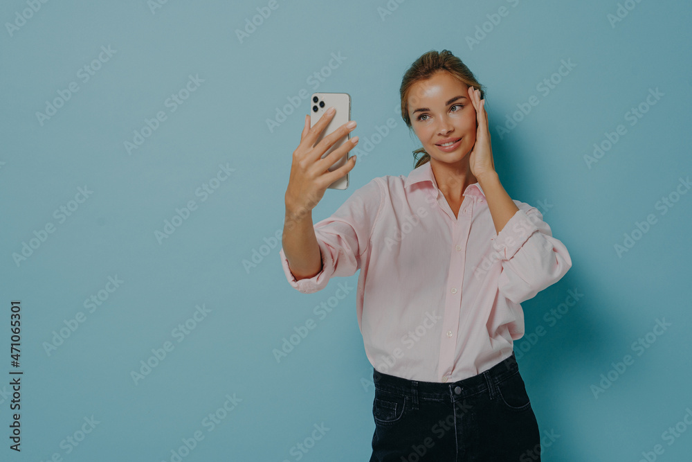 Elegant young lady in casual wear trying to take selfie while holding smartphone, isolated on blue Elegant young lady in casual wear trying to take selfie while holding smartphone, isolated on blue